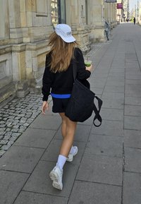 Avslappnad outfit med en svart sweatshirt, blå detaljer och svarta shorts. Bär en vit keps och har en kviltad svart väska. Vita sneakers.