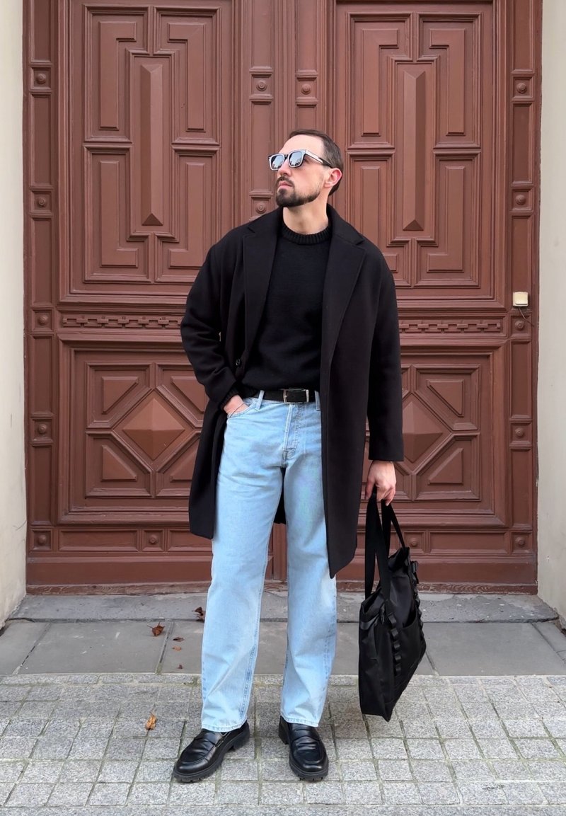 Manteau noir, pull noir, jean bleu clair, chaussures noires. Tenant un sac noir. Debout devant une porte marron texturée.