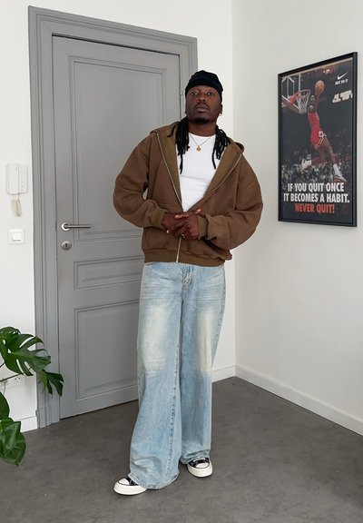 Hombre con sudadera marrón con capucha, camisa blanca, vaqueros anchos azul claro y cubierta de cabeza negra, permanece en el interior cerca de una puerta gris y un cartel motivacional de baloncesto.