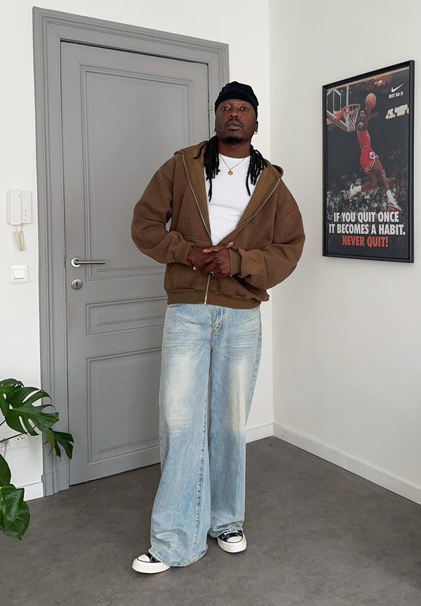 Uomo che indossa una felpa marrone con cappuccio, camicia bianca, jeans larghi azzurri e copricapo nero, in piedi all'interno vicino a una porta grigia e a un poster motivazionale di basket.