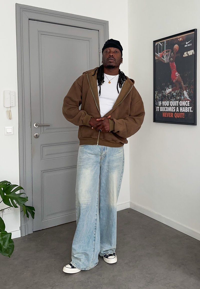 Mann trägt braunen Hoodie, weißes Hemd, hellblaue weite Jeans und schwarze Kopfbedeckung, steht drinnen nahe einer grauen Tür und einem motivierenden Basketballposter.