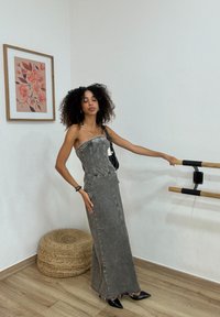 Graues Jeans-Maxikleid mit tailliertem Oberteil, seitlichen Schlitzen und dezenten Nähten. Getragen mit schwarzen spitzen High Heels. Enthält eine kleine schwarze Tasche.