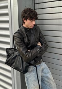 Ung mand med krøllet hår iført en sort læderjakke og lyse jeans, bærende på en stor sort lædertaske, stående op ad en metalvæg.