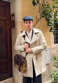 Bež trench kaput s širokim reverima, tamne hlače i smeđa kožna torba preko ramena. Siva ravna kapa. Urbani ambijent s zelenilom.