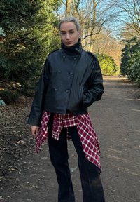 Personne debout sur un sentier en terre dans un parc, portant une veste en cuir noire, un pantalon noir et une chemise à carreaux rouges nouée autour de la taille.