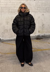 Fekete puffernadrág magas gallérral és steppelt mintával, laza fekete nadrággal és lapos fekete cipővel párosítva. Simább textúra, minimális díszítések.