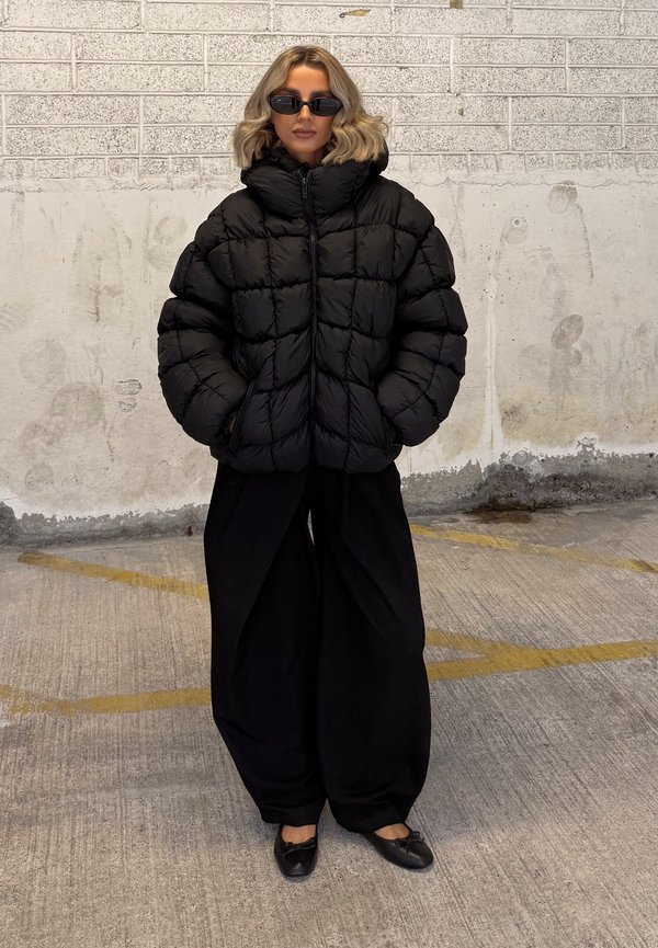 Jachetă puffer neagră cu guler înalt și design matlasat, asortată cu pantaloni negri largi și pantofi negri cu talpă plată. Textură netedă, accente minime.