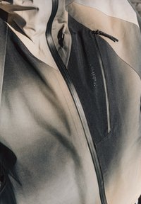 Veste résistante à l'eau avec un design ombré, présentant des tons noirs et gris, une fermeture éclair proéminente et une petite poche sur le devant à gauche.