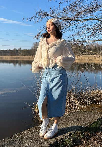 Blusa texturizada beige claro con puños rizados y un lazo, combinada con una falda de mezclilla de longitud media y zapatillas deportivas blancas.