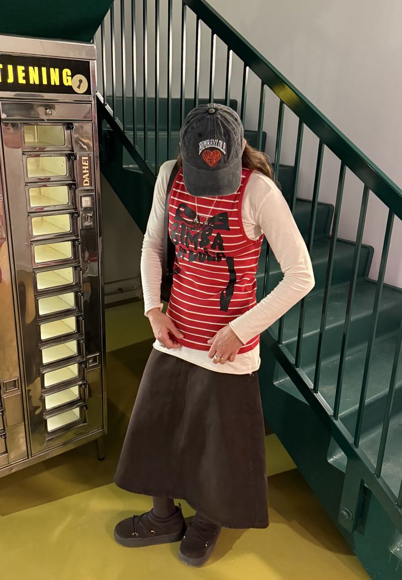 Άτομο που φοράει κόκκινη ριγέ φανέλα πάνω από λευκό μακρυμάνικο πουκάμισο και σκούρα φούστα, στέκεται κοντά σε αυτόματο πωλητή και σκάλες.