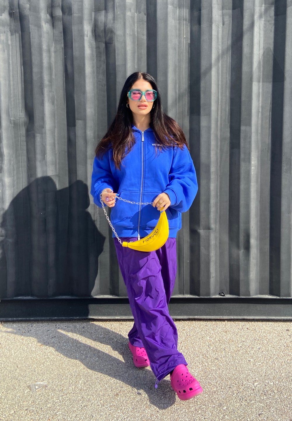 Fato de capuz azul com fecho, calças roxas, Crocs cor-de-rosa e uma bolsa em forma de banana amarela com uma corrente. O fundo apresenta linhas verticais cinzas.