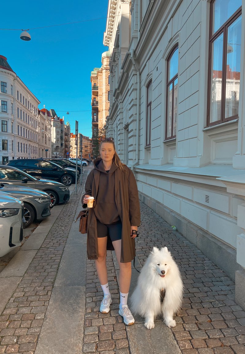 Persoon in een lange bruine jas, zwarte shorts en witte sneakers, met een drankje in de hand, naast een donzige witte Samojede op een kinderkopjesstraat.