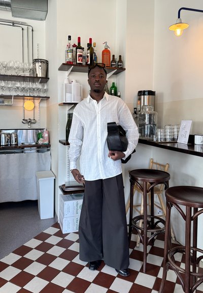 Hombre con camisa blanca abotonada y pantalones negros anchos sostiene una bolsa negra, de pie en una cafetería con estantes con botellas, cristalería y taburetes cerca.