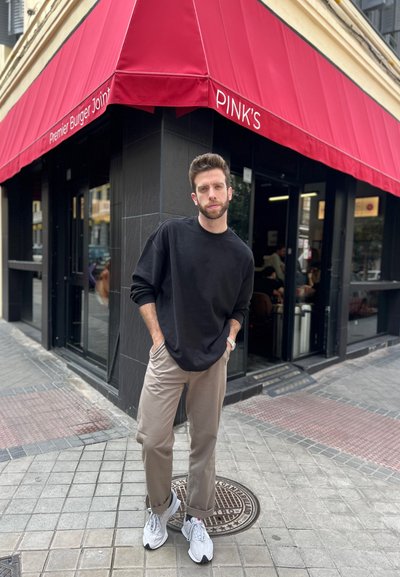 Un joven con un sudadera negra de gran tamaño, pantalones beige y zapatillas blancas está de pie frente a un restaurante con un toldo rojo.