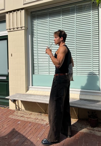 Pantalones negros holgados con bolsillos, una camiseta sin mangas negra ajustada y zapatos negros. Sosteniendo una bebida, de pie cerca de una ventana con persianas.