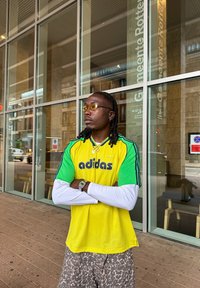 Žlutý a zelený síťovaný dres Adidas s logem, spárovaný s vzorovanými šortkami. Příslušenství zahrnuje žluté sluneční brýle a stříbrné hodinky.
