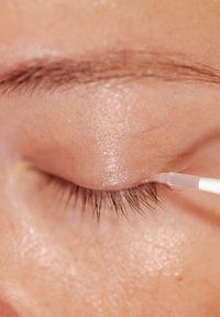 Närbild av ett öga där en klar applikationsborste applicerar en glansig produkt på ögonlocket, vilket framhäver fina fransar och hudens textur.