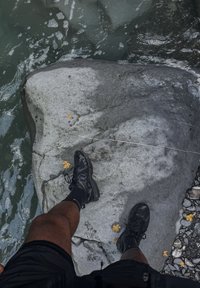 Zwarte sportschoenen met een gestructureerd ontwerp en rubberen zolen, geplaatst op een grote grijze rots nabij water met verspreide kleine gele blaadjes.