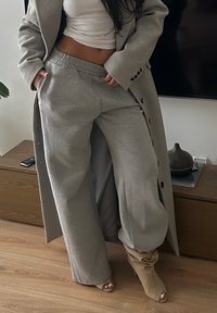 Kvinna som bär grå vida sweatpants, taupe öppna stövlar, vit kort topp och en lång grå kappa, stående inomhus nära möbler i trä.