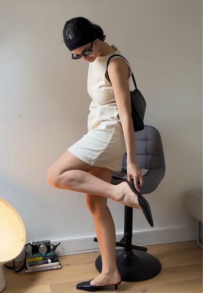 Vestido mini texturizado color blanco roto con un cuello alto, mules planos negros y un bolso negro de hombro, todo sobre un fondo neutro.