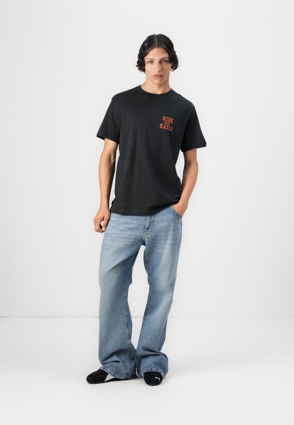 T-shirt in cotone nero con grafica rossa "RIDE THE RAILS", abbinato a pantaloni in denim larghi di colore blu chiaro e scarpe slip-on nere.