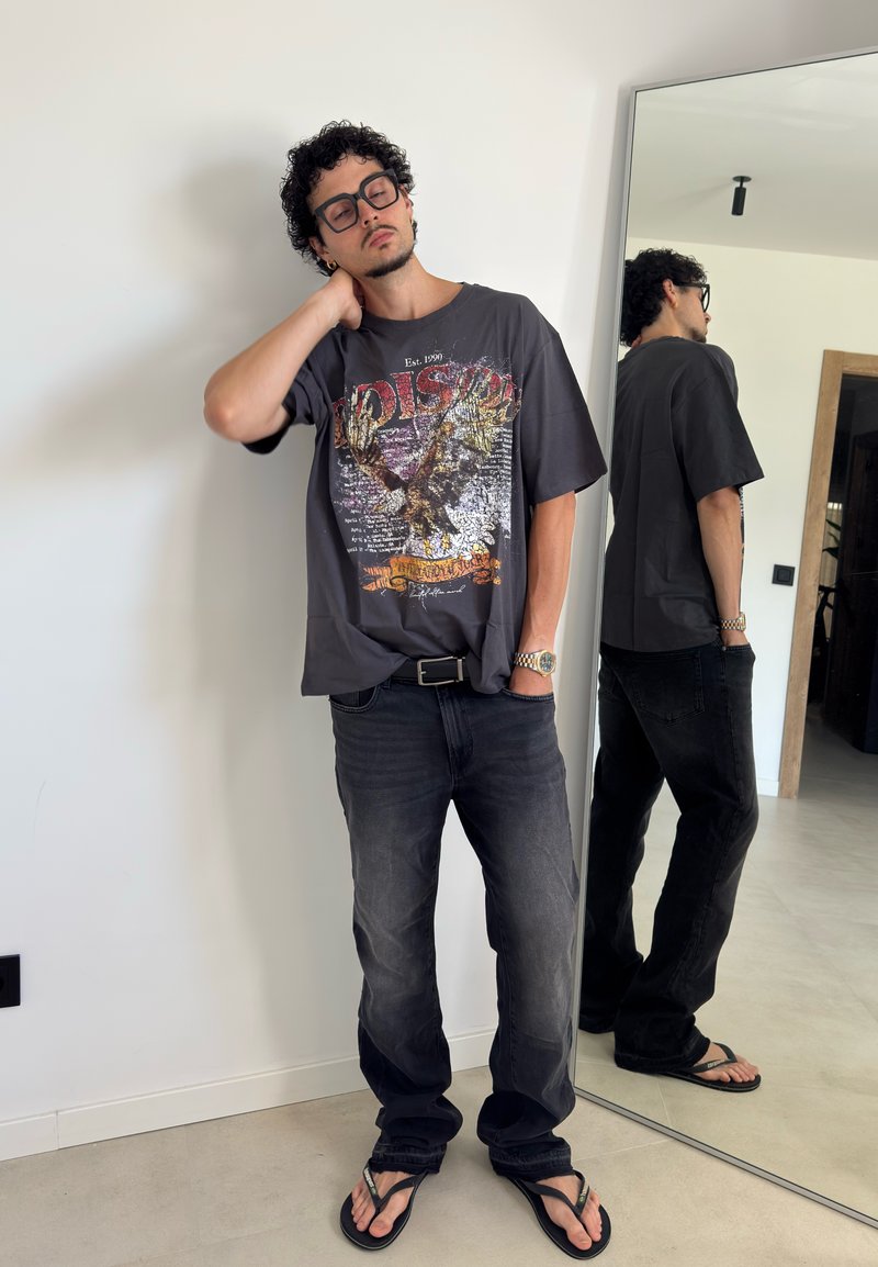 Homme avec des lunettes portant un t-shirt à motif, un jean noir et des tongs, debout avec une main sur le cou près d'un miroir en pied.