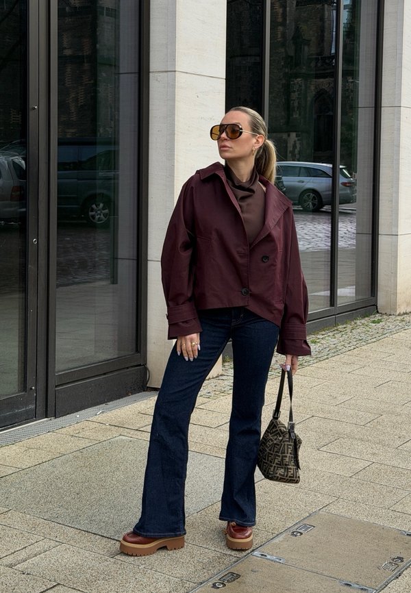 Nainen ylisuurissa viininpunaisissa takissa, tummissa farkuissa, massiivisissa ruskeissa kengissä ja aurinkolaseissa, pitää kuvioitua käsilaukkua ja seisoo kaupunkikadulla.