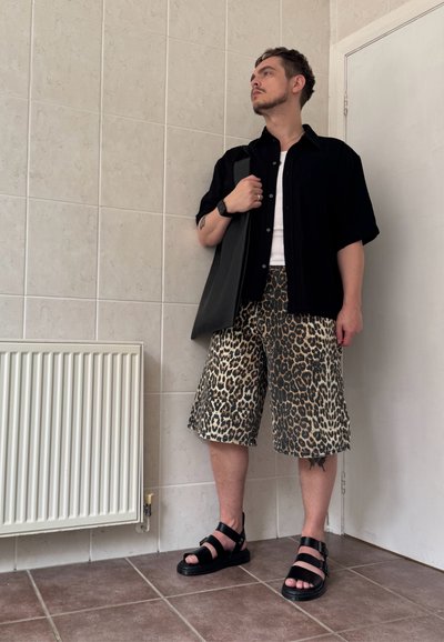 Camisa negra de manga corta sobre una camiseta blanca, combinada con pantalones cortos de estampado de leopardo. Lleva sandalias negras y lleva un bolso tote negro.
