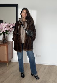 Bruine oversized jas van imitatiebont, beige hoodie eronder, blauwe rechte jeans, zwarte heuptasje en zwarte loafers met hakken, met bloemen op de achtergrond.