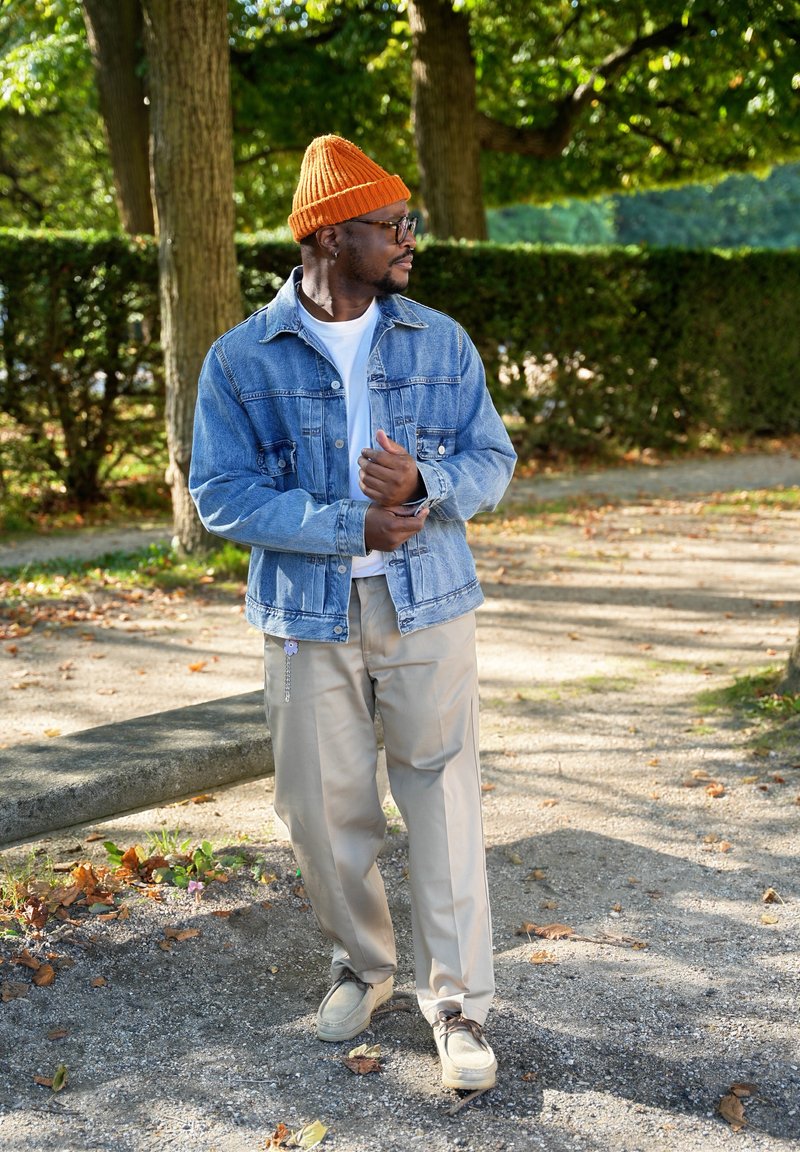 Jaqueta de denim, camisa branca, calças beiges e um gorro de malha laranja. Em pé num caminho de gravilha com folhagem verde ao fundo.