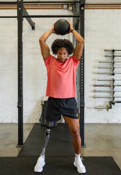 Atleta con una camiseta rosa y pantalones cortos negros levanta una balón medicinal negro texturizado por encima de su cabeza. Presenta una pierna protésica y zapatos deportivos blancos.