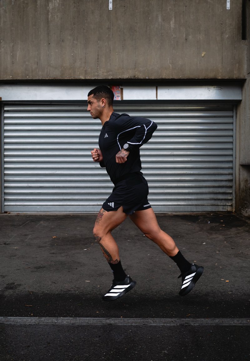 Homme athlétique avec des tatouages faisant du jogging en tenue de sport noire et chaussures de course sur un trottoir urbain devant une porte de garage en métal fermée.
