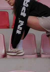 Chaussures de sport avec dessus beige, accents violets et semelle texturée. T-shirt à manches longues noir et shorts clairs. Sièges roses de stade en arrière-plan.
