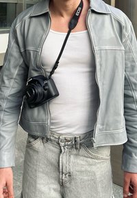 Ανοιχτό γκρίζο δερμάτινο τζάκετ με φερμουάρ, φορεμένο πάνω από λευκό ριμπ μπλουζάκι. Μαύρη φωτογραφική μηχανή Canon κρέμεται από λουρί γύρω από τον λαιμό.