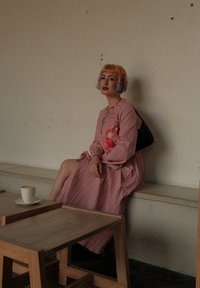 Vestido midi de color rosa con pliegues, de manga larga y un lazo en el cuello, combinado con un pequeño bolso negro y un accesorio de peluche rosa.