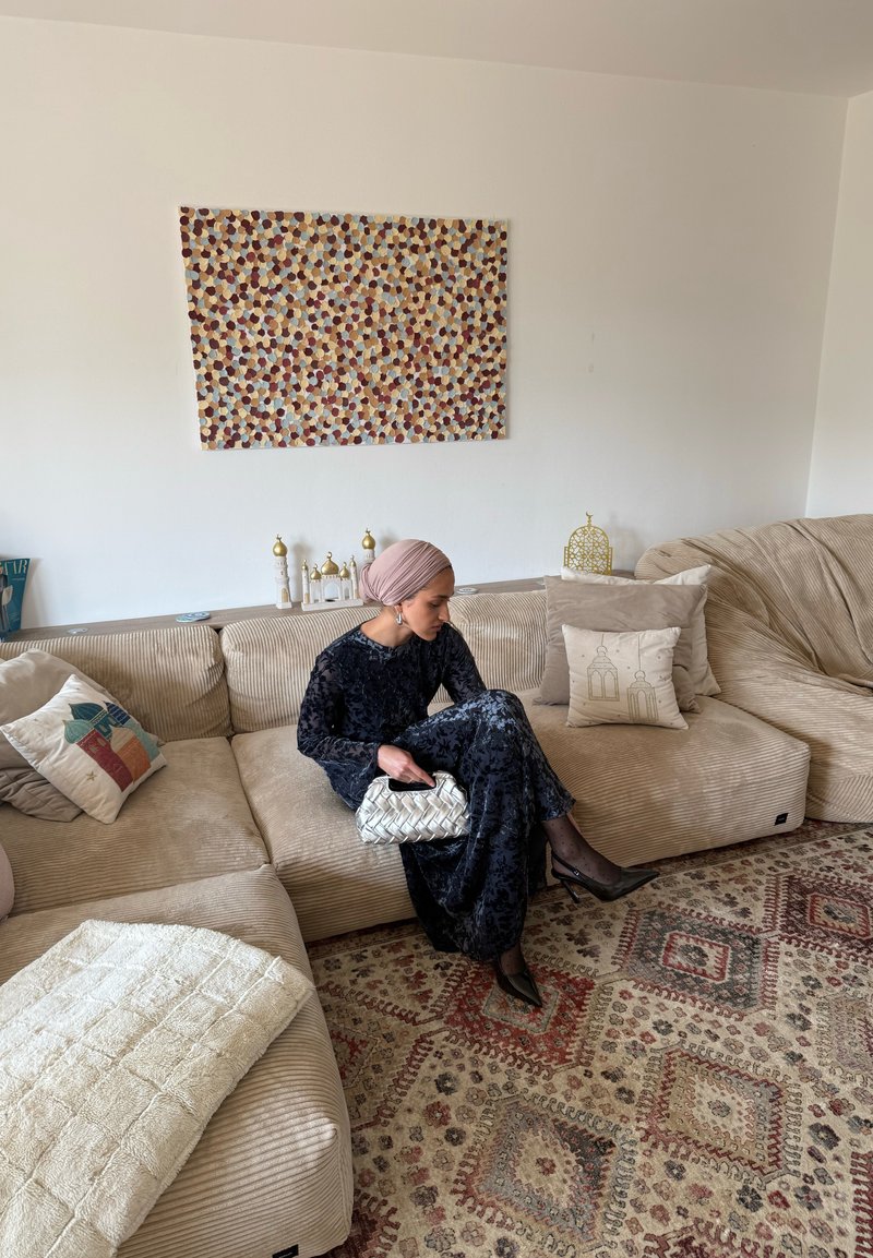 Frau in dunklem Kleid und pinkem Kopftuch sitzt auf einem beigen Sofa und hält eine weiße, geflochtene Handtasche in einem dekorierten Wohnzimmer mit gemustertem Teppich und Kissen.