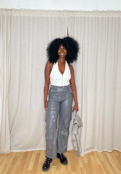 Top blanco de halter con cremallera, pantalones cargo gris de imitación de cuero con bolsillos y botas negras con cordones. Sosteniendo un bolso gris.