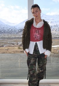 Jaqueta castanha felpuda, top de alças vermelho com design de polvo, camisa branca, calças cargo camufladas, e botas pretas. Fundo de montanha visível.