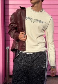 Beige geribd shirt met lange mouwen en een grijs grafisch ontwerp, gecombineerd met een bordeauxrode leren jas en zwarte sweatpants met luipaardprint en witte strepen.