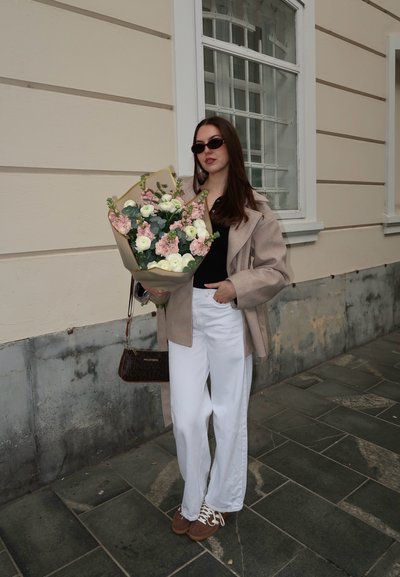 Mujer con abrigo beige, camiseta negra, pantalones blancos y gafas de sol sostiene un ramo de flores blancas y rosas mientras está de pie en una acera pavimentada.