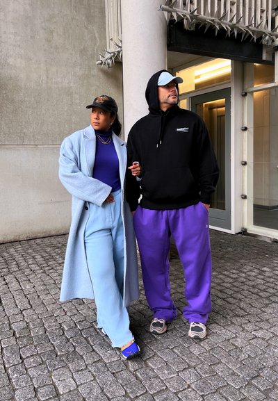 Mujer con abrigo y pantalones azul claro que enlaza el brazo con un hombre en sudadera negra, pantalones morados y gorra azul claro en una calle de adoquines.