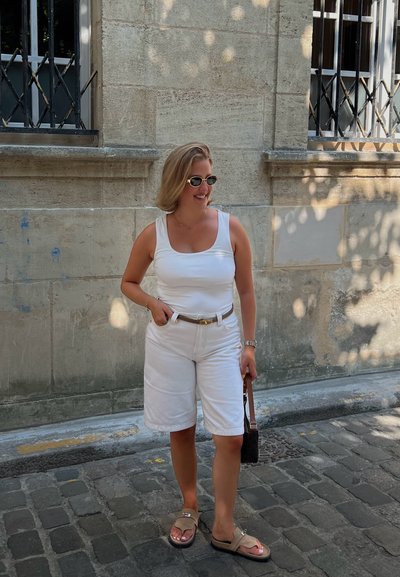 Top blanco sin mangas, pantalones cortos blancos con un cinturón y sandalias marrones. Accesorios incluyen gafas de sol redondas y un bolso pequeño. Ambiente casual al aire libre.