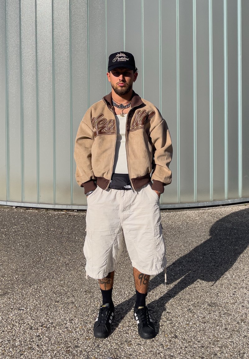 Homme avec barbe et casquette portant une veste beige, une chemise blanche, un short cargo clair, des chaussettes noires et des baskets, debout sur du gravier devant un mur en métal.