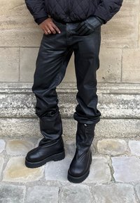 Bottes montantes noires en cuir avec une semelle épaisse, portées avec un pantalon large en cuir noir et une veste matelassée noire. Matériaux texturés.