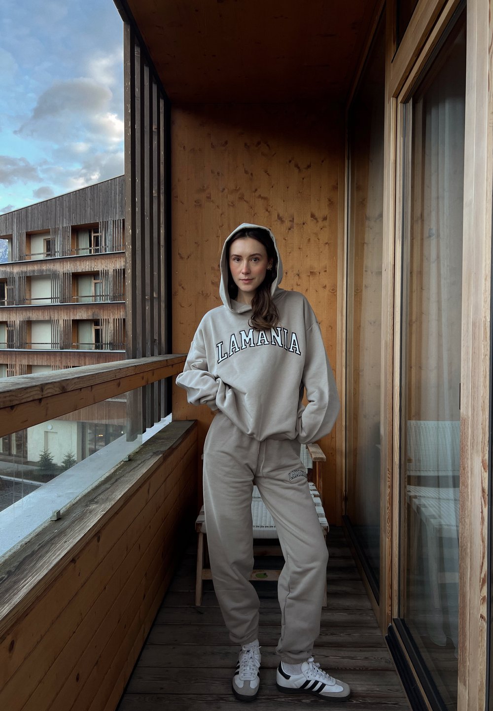 Sweat à capuche gris clair avec le texte "LAMANIA", jogging assorti et baskets blanches. Balcon en bois avec vue sur des bâtiments modernes. Fond en bois texturé.