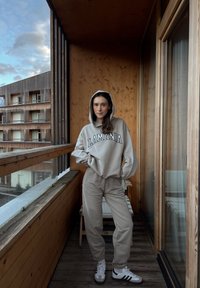 Sweat à capuche gris clair avec le texte "LAMANIA", jogging assorti et baskets blanches. Balcon en bois avec vue sur des bâtiments modernes. Fond en bois texturé.