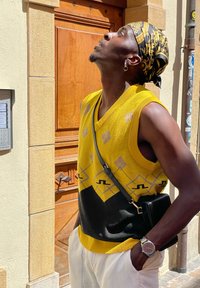 Αμάνικο κίτρινο και μαύρο πουλόβερ με γεωμετρικά σχέδια, συνδυασμένο με μαύρη τσάντα χιαστί, φορεμένο με λευκά σορτς.