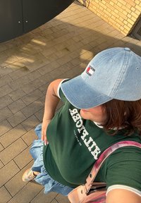 Πράσινο πουκάμισο με λευκό κείμενο, κοντή τζιν φούστα, ανοιχτό μπλε καπέλο με λογότυπο, ροζ τσάντα ώμου, καφέ αθλητικά παπούτσια και υφή στο έδαφος.