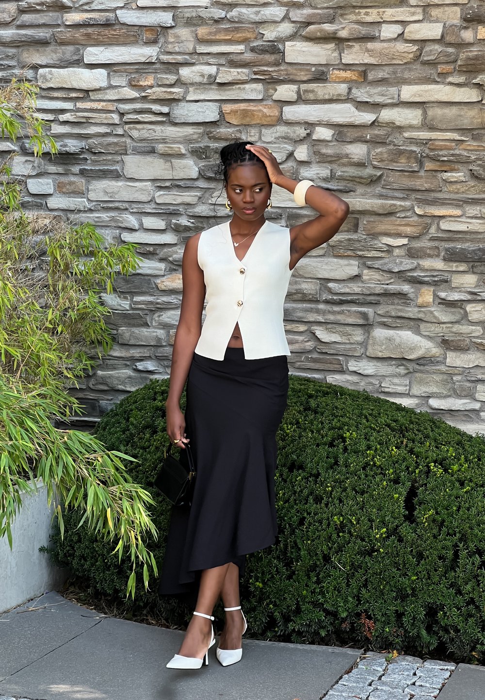 Κρεμ αμάνικο τοπ με V-λαιμόκοψη και χρυσά κουμπιά, συνδυασμένο με μια μαύρη μίντι φούστα. Λευκά πέδι�λα με λουράκι στον αστράγαλο και μια μικρή μαύρη τσάντα.