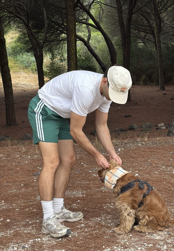 Uomo con t-shirt bianca e cappellino si piega per sistemare un foulard sulla testa di un cane marrone soffice seduto in un'area esterna boscosa.